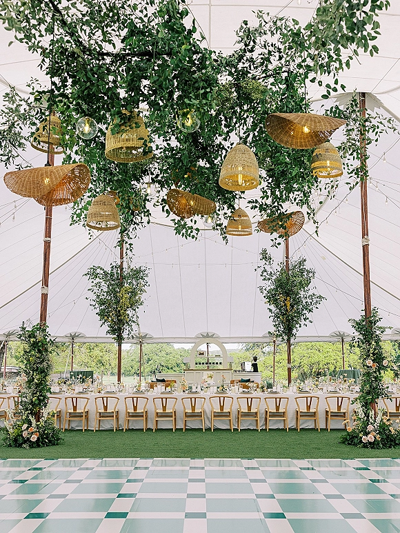 Reception decor with a tented wedding reception featuring a long banquet table, greenery canopy, wicker pendants, string lights, and candles under a white tent