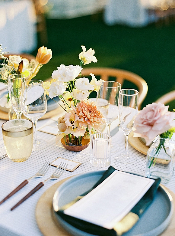 Reception tablescape with wedding place setting, gold charger plates, floral centerpiece, bud vases, mixed glassware and votive candles on an outdoor lawn