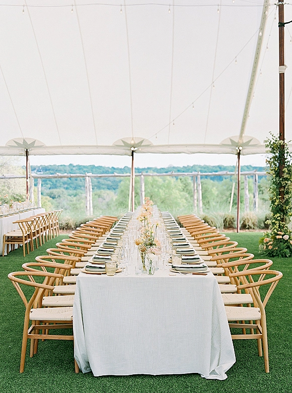 Reception tablescape on a long banquet table wedding under a tent canopy, with pastel bud vases, taper candles, and string lights over rolling hills