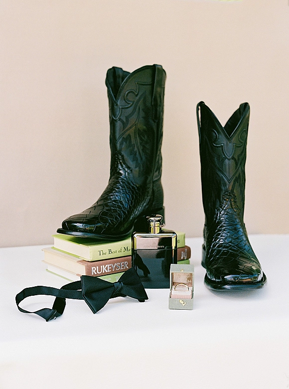 Groom accessories flat lay with black cowboy boots, bow tie, cologne, wedding rings and ring box on stacked books on a white tabletop