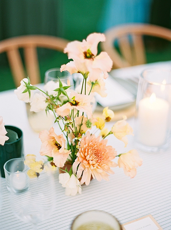 Wedding centerpiece with pastel floral centerpiece of peach and white blooms, greenery, and glass hurricane candle on a striped tablecloth outdoors