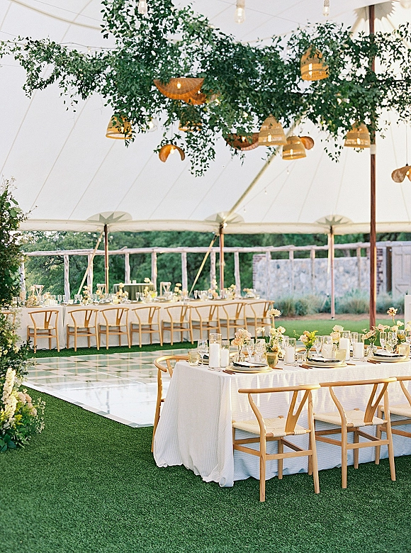 Reception tablescape with white linens, wishbone chairs, taper candles and small florals under hanging greenery in a tent with string lights