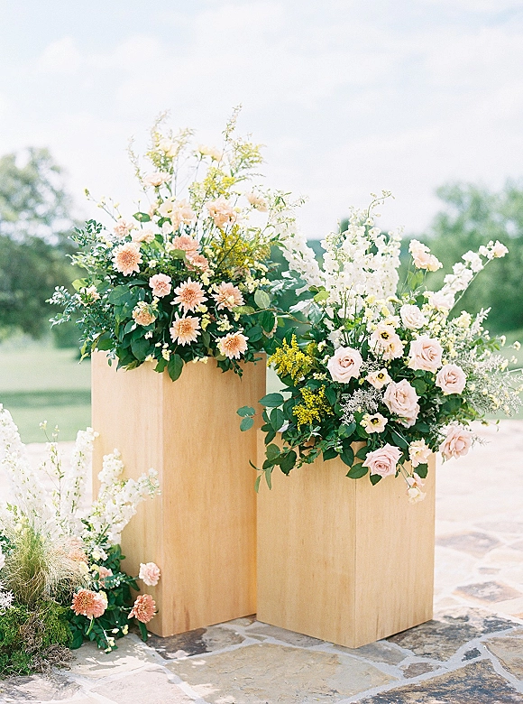 Wedding floral pedestals with ceremony pedestal flowers on wood stands, featuring blush roses and peach dahlias on a stone patio lawn backdrop
