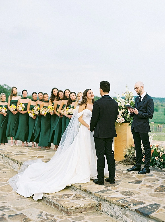 Wedding ceremony moment as bride in strapless gown and long veil holds groom’s hands while officiant reads on a stone patio with hills beyond
