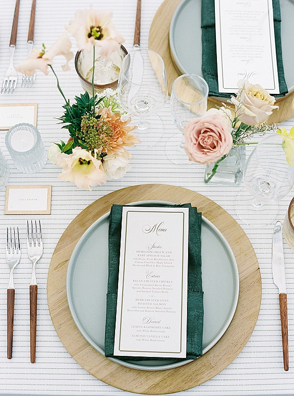 Reception tablescape with wedding place setting featuring menu card on a dark green napkin, sage plate, wood charger, rose centerpiece, votive candle on striped linen