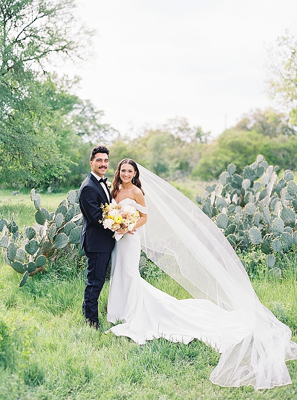 Couple portrait of bride holding a pastel bouquet beside groom in tuxedo, long veil blowing in wind with cactus plants behind them