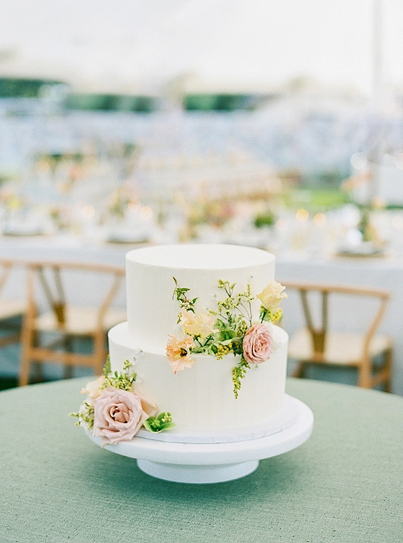 Wedding cake with blush roses and peach flowers on a cake stand, white buttercream tiers set by rustic outdoor reception tables