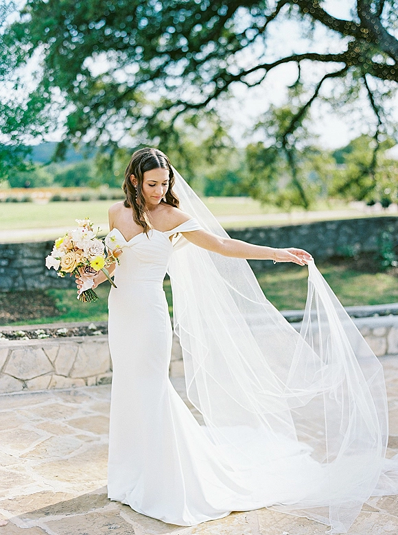 Bridal portrait of a bride holding bouquet, wearing a strapless wedding dress and long veil on a stone patio with trees and sky behind
