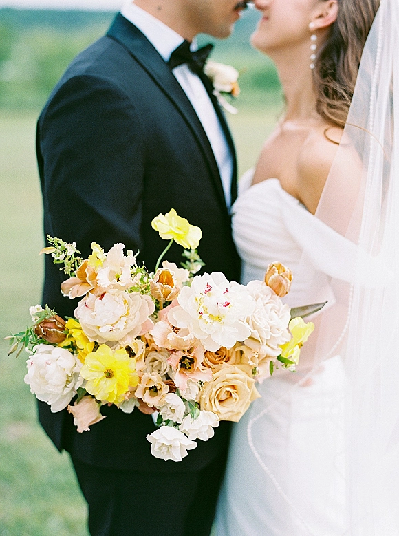 Wedding couple portrait of bride and groom holding bouquet, leaning in for a close-up kiss with veil and tuxedo amid outdoor greenery