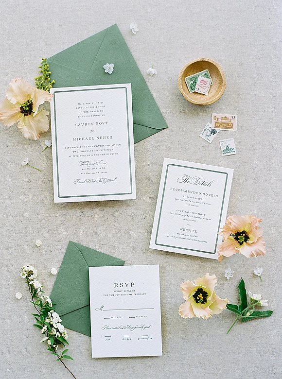 Invitation suite with a green envelope, RSVP and details cards, and postage stamps styled with flowers and greenery on linen fabric