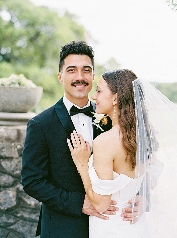 Couple portrait of bride and groom embrace, bride looking at groom under a veil with pearl earrings by a stone wall and greenery