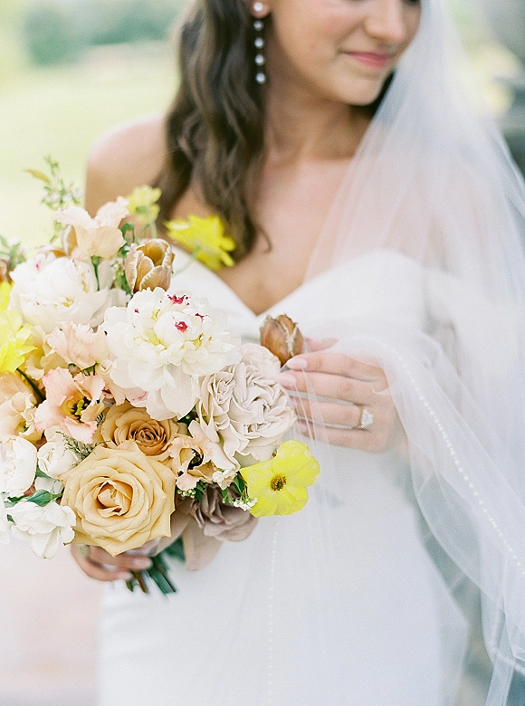 Bridal bouquet of roses, peonies, and ranunculus with greenery and ribbon wrap held by bride in strapless dress outdoors in soft blur