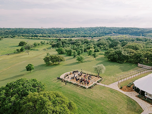 Outdoor wedding ceremony shown in an aerial wedding ceremony view, with circular seating, aisle, and floral accents in a hilltop field near a tent