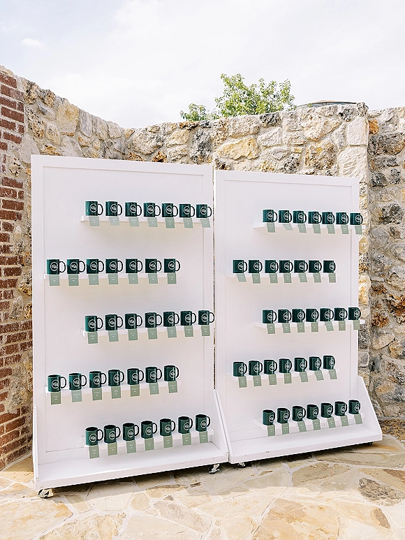 Wedding favor display with personalized wedding favors mugs and place cards on white shelves, set against a stone and brick patio wall outdoors