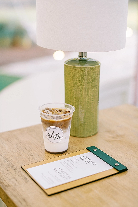 Wedding drink favor iced coffee in a clear plastic cup with custom monogram sticker beside a coffee menu card on a wooden table, soft lights behind