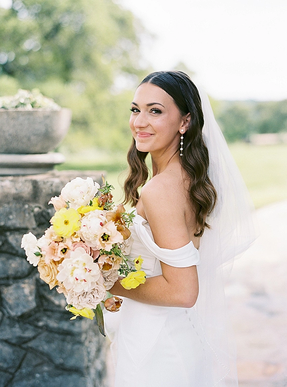 Bridal portrait of a smiling bride holding bouquet, looking over her shoulder in an off the shoulder dress with veil by a stone fountain