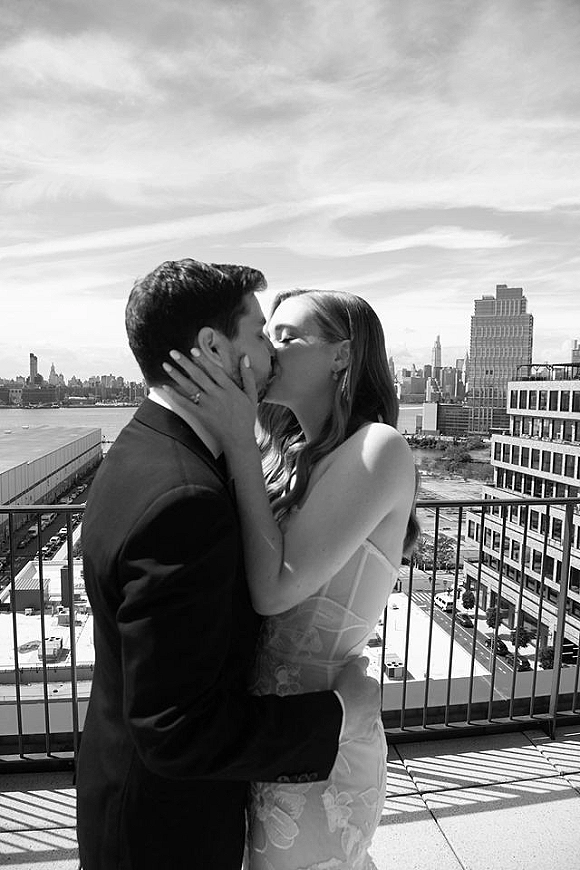 Wedding kiss portrait in black and white as bride in lace gown and groom in suit embrace on a rooftop terrace with city skyline behind