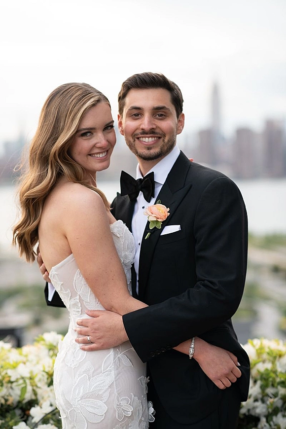 Couple portrait of bride and groom hugging and smiling, her strapless lace dress beside his tux, on a terrace by water and skyline