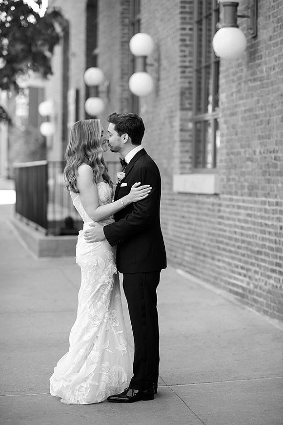 Wedding couple portrait of bride and groom kiss in close embrace, strapless lace dress and black tuxedo by brick wall on city sidewalk