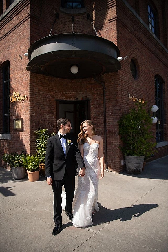 Couple portrait of bride and groom walking hand in hand, her strapless lace dress and his black tuxedo by a brick doorway under an awning