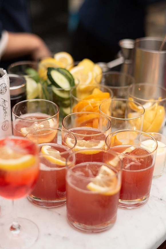 Wedding signature drinks on a signature cocktail bar with citrus and cucumber garnishes, cocktail glasses on a marble top with guests blurred behind