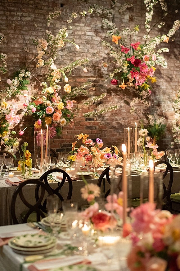 Reception tablescape with wedding table decor, colorful floral centerpieces, taper candles in glass hurricanes, and black chairs against exposed brick wall