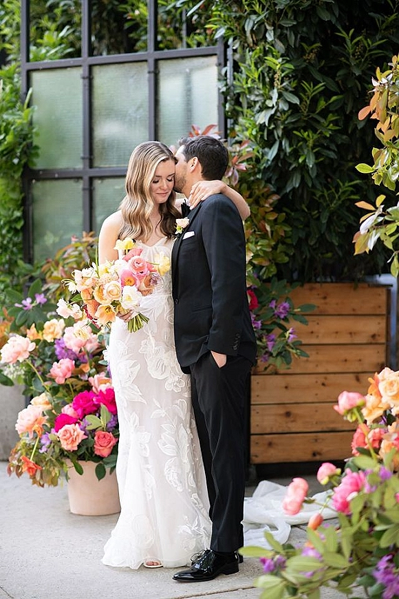 Couple portrait of bride and groom embrace with a forehead kiss, bouquet accent, framed by greenhouse windows and a greenery wall