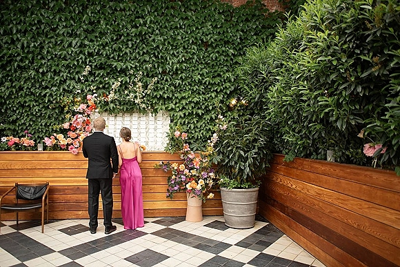Wedding bar setup with colorful floral arrangements on a wood slat counter, against a white tile backdrop and ivy wall on a checkered floor