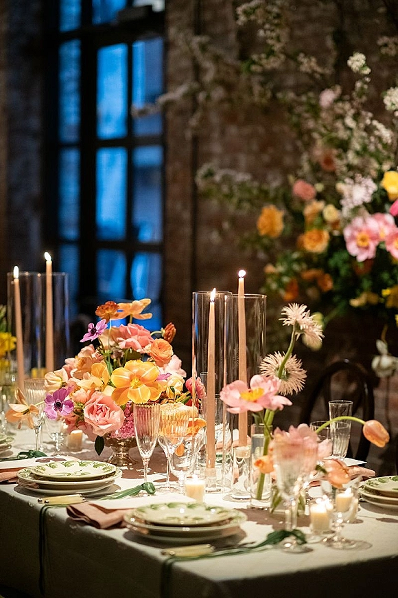 Reception tablescape with wedding table centerpiece of colorful florals and taper candles, set with patterned plates and glassware by brick wall window