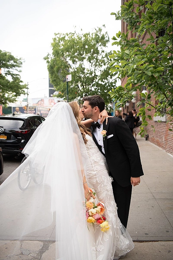 Wedding kiss portrait of bride and groom kissing on a city sidewalk, her veil blowing as she holds a colorful bouquet beside a brick wall