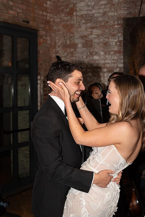First dance moment as bride and groom dance closely, her strapless lace dress and bracelet visible, guests and brick wall behind