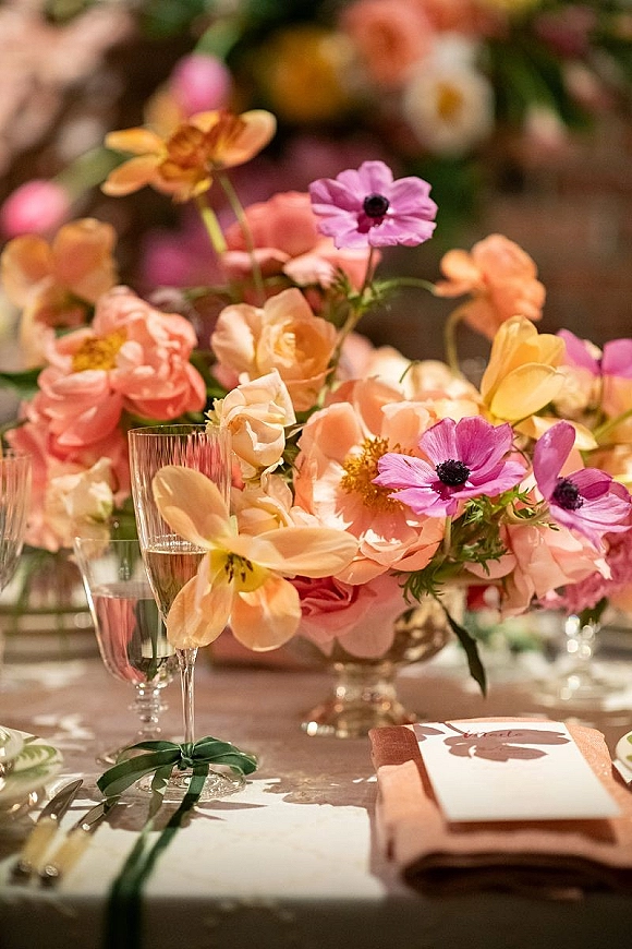 Reception tablescape with a wedding table centerpiece of lush peach and pink blooms, bud vases, champagne flutes, menu cards, and silver flatware on white linens