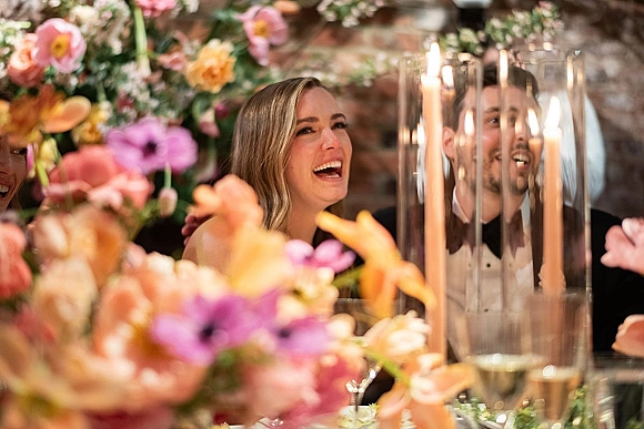 Reception candid moment of newlyweds laughing at dinner at a brick wall head table, with colorful florals and tall taper candles in glass hurricanes