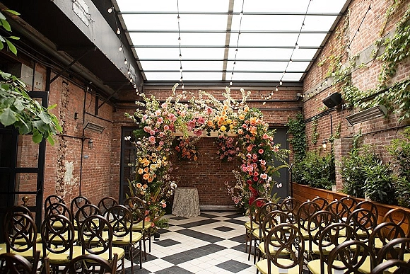 Ceremony setup with a floral ceremony arch under hanging string lights, chairs lining a checkerboard aisle in a brick-walled courtyard