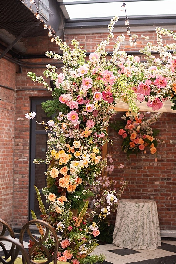 Wedding floral installation with a ceremony floral arch of pink and peach roses and greenery, set against an exposed brick wall with bistro lights