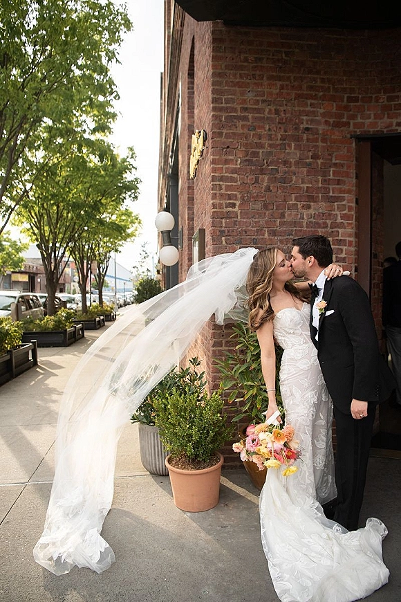 Wedding kiss portrait of bride and groom kissing as her long veil blows in the wind, leaning by a brick building with potted greenery