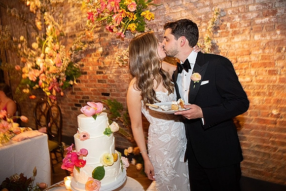 Wedding cake cutting as bride in a strapless lace dress and groom in a black tuxedo kiss by a three-tier cake against a brick wall