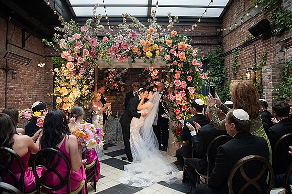 Ceremony kiss under a floral arch as the bride and groom kiss beneath a chuppah in a brick courtyard with glass ceiling and string lights