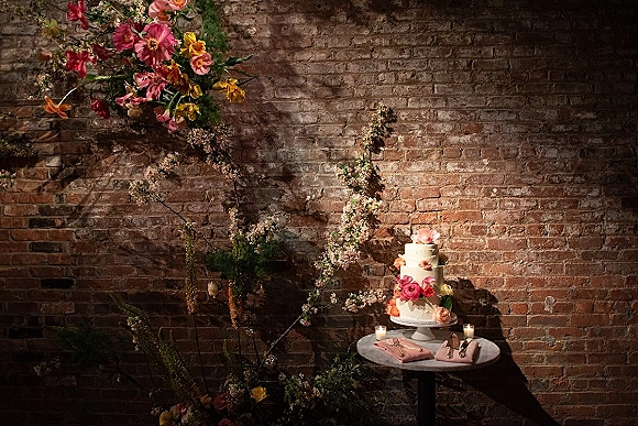 Wedding cake display featuring a tiered wedding cake with sugar flowers, greenery accents, and votive candles against an exposed brick wall