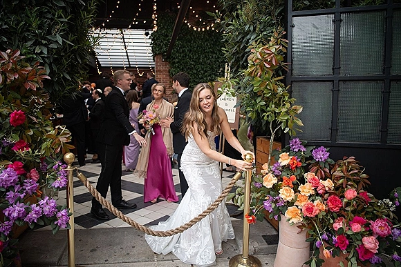 Bride entrance with rope stanchions as she opens the barrier in a courtyard, guests mingling near a floral welcome sign and string lights