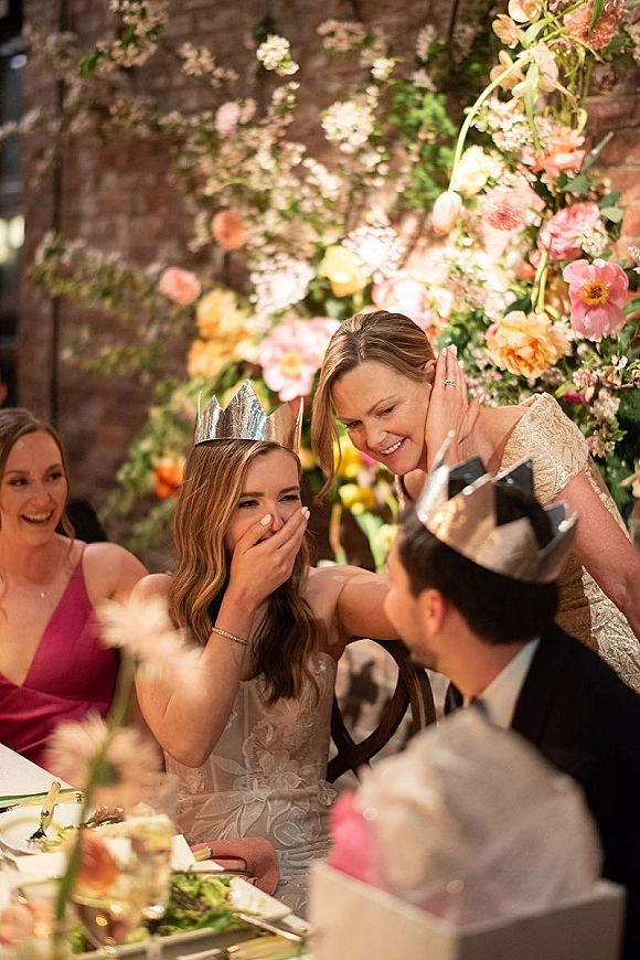 Wedding reception moment as bride laughs covering her mouth beside groom in paper crown at head table with floral backdrop and brick wall