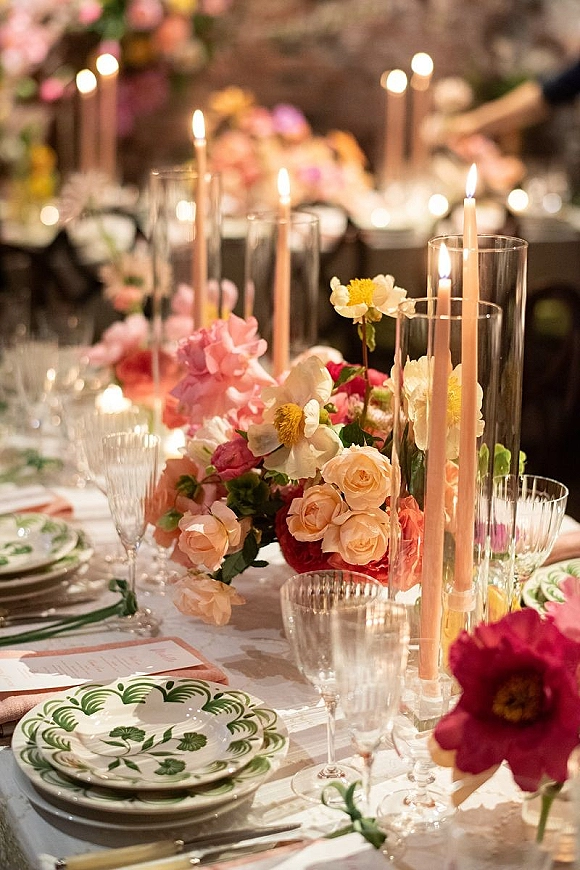 Reception tablescape with wedding table centerpiece of pink and peach florals, taper candles, crystal stemware, and patterned plates on a warm-lit banquet table