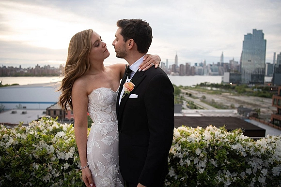 Wedding kiss portrait of bride and groom kissing on a rooftop terrace, strapless lace gown and tux, with city skyline and river behind