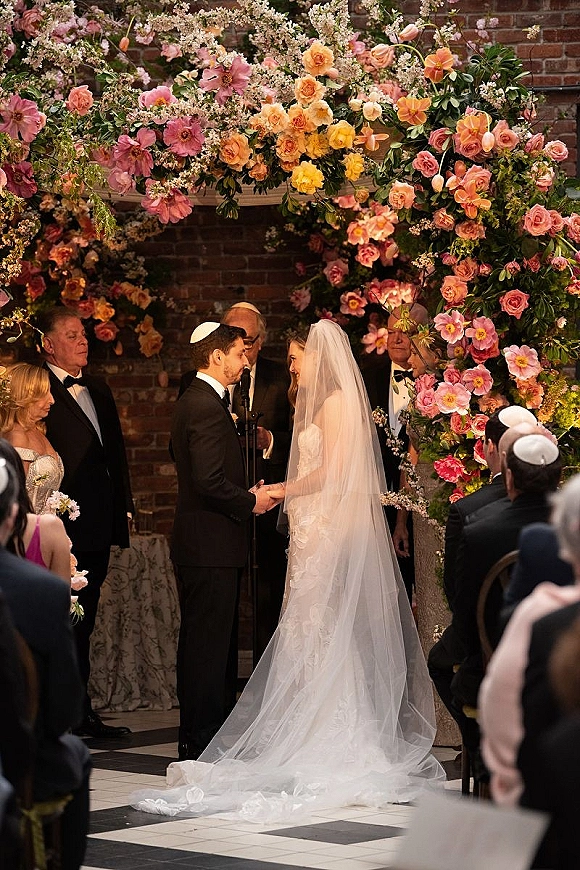 Wedding vows as bride and groom hold hands under a rose floral arch, veil and long train flowing in an indoor brick-walled ceremony space