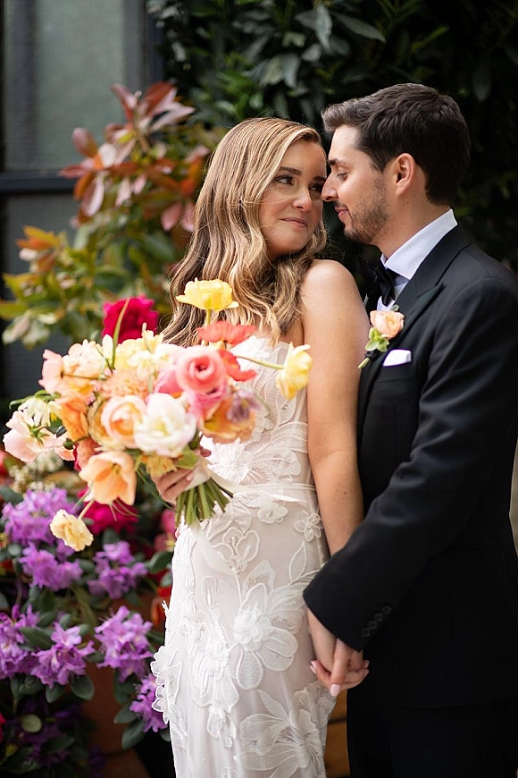 Couple portrait of bride and groom holding hands, her colorful bridal bouquet and lace dress beside his tux, amid lush garden foliage near a window
