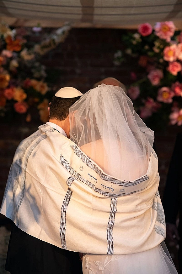 Ceremony moment at a Jewish wedding ceremony as bride and groom embrace from behind under a chuppah, veil and tallit with floral arch greenery