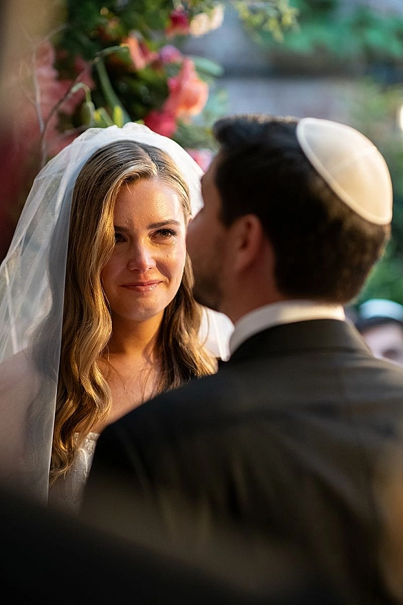 Ceremony moment as bride in veil listens to wedding vows from groom in suit and kippah, tearful smile amid outdoor greenery and guests