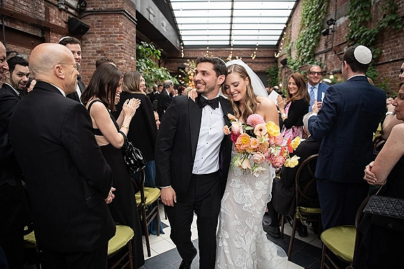 Wedding recessional as bride and groom walk the aisle, bride in lace dress and veil with bouquet, guests clapping under string lights in brick loft