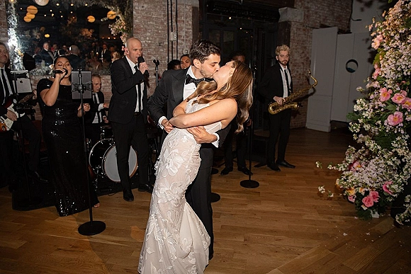 Wedding kiss as groom dips bride in strapless lace dress on wooden dance floor, live band and floral wall glowing under hanging lights