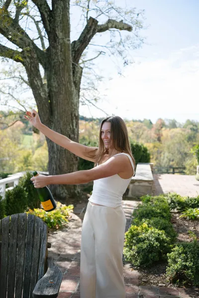 Champagne pop as a woman in a white camisole and cream wide-leg pants sprays a champagne bottle, engagement ring visible on a garden patio